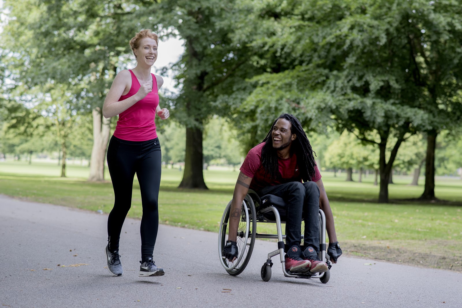 A woman in a pink t-shirt running alongside a man in a wheelchair through a park. Both are laughing.